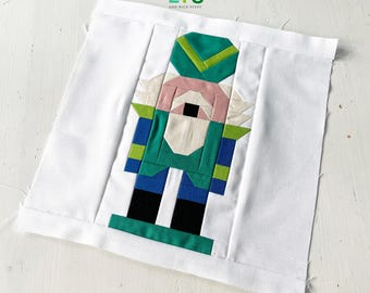 May include: A white fabric square with a green, blue, black, and pink patchwork design of a nutcracker. The fabric square has the text "QUILTS AND NICE STUFF" in the top left corner.