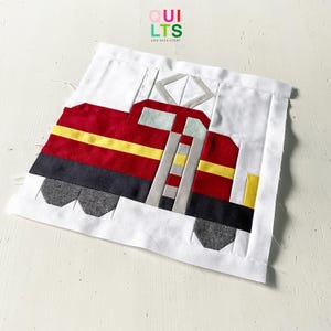 Könnte beinhalten: Ein handgefertigter Quiltblock mit einem Zugdesign in Rot, Gelb, Grau und Schwarz. Der Zug ist vor einem weißen Hintergrund platziert. Der Text "QUILTS and NICE STUFF" ist oben rechts zu sehen.