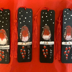 Puede incluir: Cuatro marcapáginas de madera negra con borlas rojas. Cada marcapáginas presenta un petirrojo pintado a mano posado en una rama con bayas rojas y un fondo nevado. La palabra "LOVE" está escrita en cada marcapáginas.