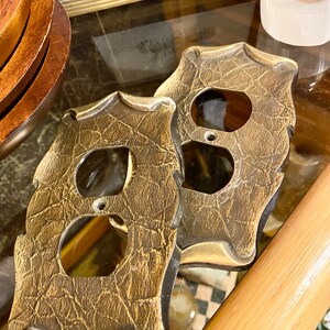 May include: Two gold-toned outlet covers with a textured surface. The covers have a decorative, ornate design.