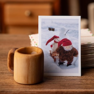 May include: A small, light-coloured wooden mug with a curved handle sits next to a rectangular card featuring a printed image of a white creature and a brown, fuzzy figure, both wearing red pointed hats, standing in the snow.