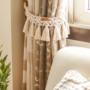May include: A close-up of a beige and brown patterned curtain with a white macrame curtain tie-back with tassels. The tie-back has a brown leather tag with the text "Boho by Brand" on it.