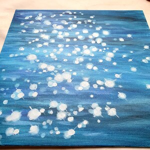 May include: Square canvas art with an abstract water scene. The painting features a gradient of blues, with white, circular highlights suggesting light reflecting on the water's surface. The artwork is on a textured canvas.