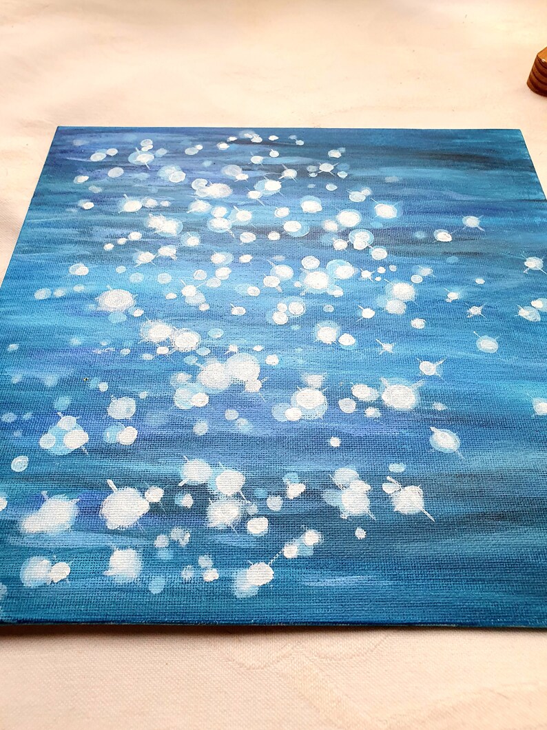 May include: Square canvas art with an abstract water scene. The painting features a gradient of blues, with white, circular highlights suggesting light reflecting on the water's surface. The artwork is on a textured canvas.