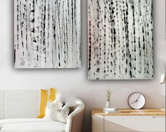 May include: Two abstract paintings in black and white with a white background and black vertical lines. The paintings are hanging on a white wall above a white couch with yellow pillows. There is a wooden table with two drawers and a clock on top of the table.