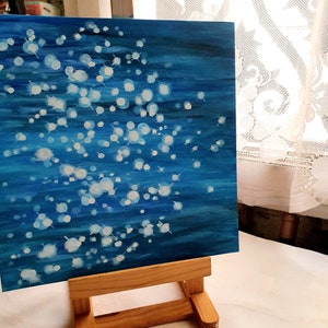 May include: An abstract painting on canvas, featuring a deep blue background with white, circular brushstrokes. The artwork is displayed on a small wooden easel, creating a gallery-like presentation. The painting evokes a sense of depth and movement.