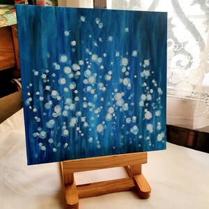 May include: An abstract painting on a square canvas, featuring a deep blue background with white and light blue circular shapes. The artwork is displayed on a small wooden easel, showcasing the painting's texture and color variations. The painting evokes a sense of depth and movement.
