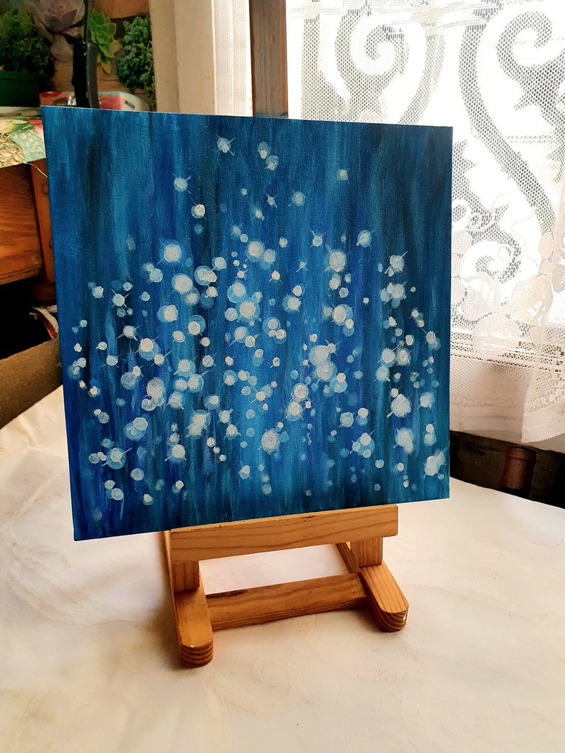 May include: An abstract painting on a square canvas, featuring a deep blue background with white and light blue circular shapes. The artwork is displayed on a small wooden easel, showcasing the painting's texture and color variations. The painting evokes a sense of depth and movement.