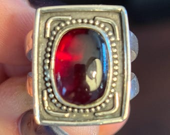 Peut inclure: Bague argentée avec un sertissage rectangulaire. Elle présente une pierre précieuse ovale rouge foncé, entourée d'une bordure perlée et d'un cadre décoratif. Le corps de la bague est divisé et présente un motif texturé.