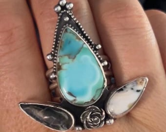 Peut inclure: Une bague en argent avec une pierre turquoise, une pierre blanche et une pierre noire. La bague est sertie dans un motif floral.
