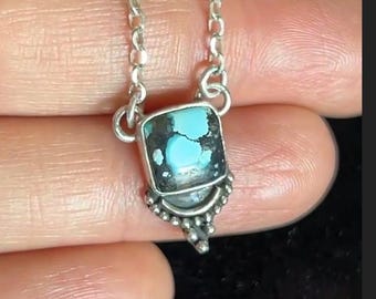 Peut inclure: Un collier en argent avec un pendentif en pierre turquoise de forme carrée présentant des inclusions de matrice sombre. Le pendentif est serti dans une monture en argent avec une base perlée et suspendu à une fine chaîne en argent.