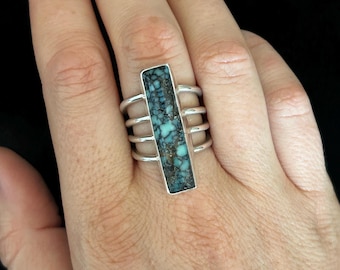 Peut inclure: Bague en argent ornée d'une pierre turquoise rectangulaire aux nuances bleues et marron. Le design moderne de la bague présente quatre bandes parallèles de chaque côté de la pierre.