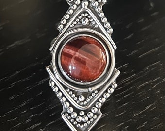 Peut inclure: Une bague en argent avec une pierre en œil de tigre rouge sertie dans un motif géométrique.