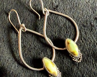 Peut inclure: Une paire de boucles d'oreilles argentées en forme de goutte d'eau. Chaque boucle d'oreille est ornée d'un pendentif ovale vert clair et marron bordé de perles argentées. Présentées sur une surface sombre texturée.
