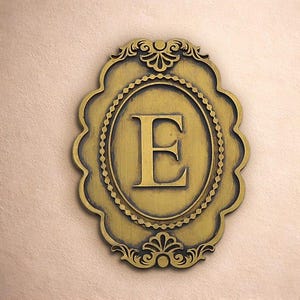 May include: A gold-colored, oval-shaped wall plaque with the letter "E" in the center. The plaque has a decorative border and ornate detailing at the top and bottom. The background is a light beige color.