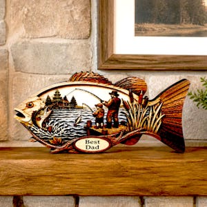 May include: Wooden fish-shaped decorative piece with a fishing scene carved inside. The scene depicts a father and child fishing, with the words "Best Dad" on an oval plaque. The piece is made of wood and has a natural finish.