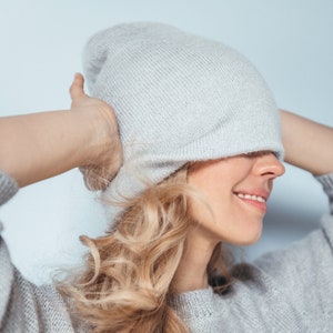 Peut inclure: Un bonnet en tricot gris clair couvrant la tête, associé à un pull assorti. Le bonnet est tiré sur les yeux. De longs cheveux blonds ondulés tombent sur le côté. Le pull a un col rond.