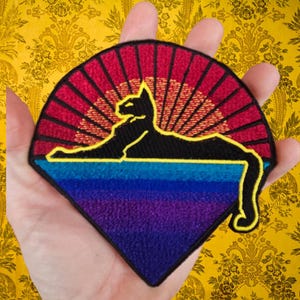 May include: Embroidered patch featuring a black cat silhouette lounging against a sunset design. The patch has a fan shape with a gradient of colors, including red, orange, blue, and purple. The cat is outlined in yellow.