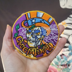Pode incluir: Remendo bordado redondo com um desenho de caveira usando uma cartola. O remendo apresenta cores roxas, laranja e azul, com o texto "GGMDead" em dourado. O remendo tem uma borda amarela e é segurado em uma mão.