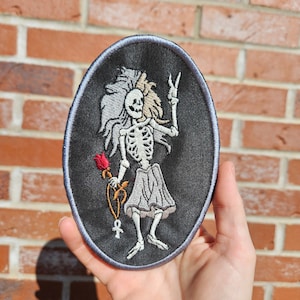 Pode incluir: Patch bordado oval com uma figura de esqueleto. O esqueleto tem cabelo esvoaçante, uma rosa vermelha e um ankh. Está fazendo o sinal de paz. O patch tem um fundo preto com uma borda azul. Design gótico.