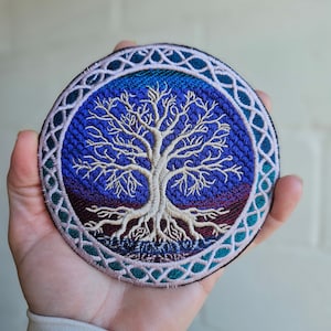 May include: Circular embroidered patch with a detailed tree design. The tree has a light beige trunk and branches, set against a deep blue and purple background. The patch is bordered by a decorative teal and beige pattern.