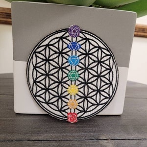 May include: A white and black embroidered patch with a flower of life design and seven colorful chakras. The chakras are arranged vertically in a line down the center of the patch.