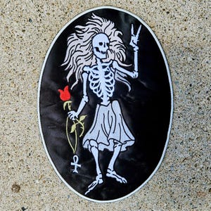 May include: Oval-shaped black patch featuring a white skeleton with flowing hair, a skirt, and a peace sign. The skeleton holds a red rose and a gold ankh. The patch has a white border.