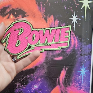 Peut inclure: Un patch brodé rose et vert avec le mot "BOWIE" dans une police de caractères stylisée.