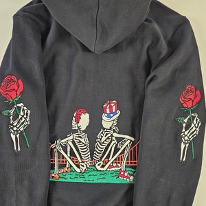 Puede incluir: Sudadera con capucha negra con un gráfico de dos esqueletos sentados en un puente, sosteniendo rosas. Los esqueletos llevan sombreros, uno con una flor roja y el otro con un sombrero rojo, blanco y azul.