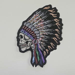 May include: Embroidered patch depicting a skull wearing a Native American headdress. The headdress is black with colourful feathers and a grey band. The skull has a detailed design with triangular shapes and a colourful pendant.