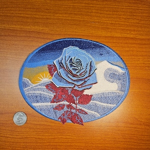 May include: An oval embroidered patch featuring a blue rose with red leaves and stem. The background depicts a sunset over a wavy ocean. The patch is approximately 7.6 cm in diameter.
