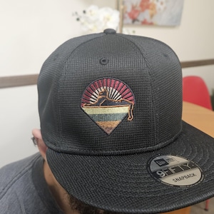 Könnte beinhalten: Schwarze Snapback-Kappe mit strukturiertem Muster. Sie hat einen gestickten Aufnäher mit einem geometrischen Design in Rot, Gold und Blau. Auf dem Schirm befindet sich ein 9FIFTY Snapback-Etikett.