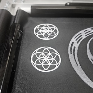 May include: Close-up of black fabric with white embroidered designs. Two circular patterns with a flower-like design are visible, along with other geometric shapes. The fabric is likely part of a larger project.
