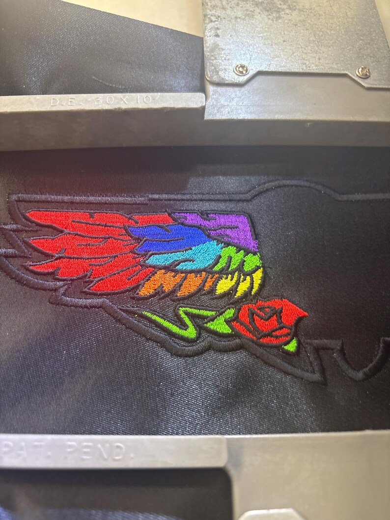 Pu&ograve; includere: Una superficie in tessuto nero con un ricamo raffigurante un'ala con piume color arcobaleno e una rosa rossa alla base. Il ricamo &egrave; fissato all'interno di un telaio metallico con il testo D.E. 80 X 10 e PAT. PEND. visibile sul metallo.