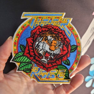 May include: A colourful embroidered patch featuring a tiger's face inside a red rose with green leaves. The patch has a blue background with a yellow border and the text "Tiger Rose" in white.