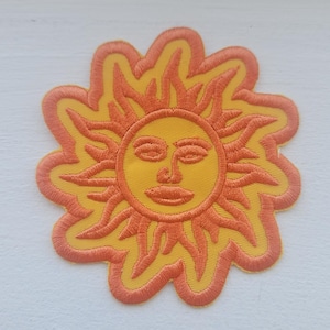 May include: Embroidered patch featuring a stylized sun with a smiling face. The sun has a yellow center and orange rays and border. The patch is likely for clothing or accessories.