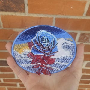 May include: An embroidered patch featuring a blue rose with red stem and leaves, set against a mountain landscape with a sunset. The patch has a blue oval border and is held in a hand.