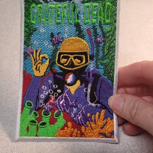 May include: Embroidered patch featuring a diver with a yellow helmet and goggles, giving an 'okay' hand gesture. The patch has the words "Grateful Dead" in green, with an underwater scene in the background. The diver is wearing a purple wetsuit.