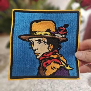 May include: Embroidered patch featuring a portrait of a person wearing a hat and scarf. The patch has a blue background with a yellow border. The person's hat is yellow and brown, and the scarf is red and orange.