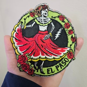 Pode incluir: Um remendo redondo bordado com um esqueleto vestido com um vestido vermelho e branco, dançando com uma borda floral verde e o texto "EL PASO".