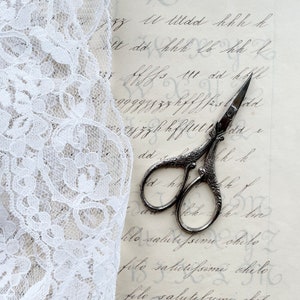 May include: A pair of antique silver scissors with ornate handles, lying on a piece of white lace fabric with a handwritten note in the background. The note contains the text "zzz U Uldd hhh h lt dd hhh lb hh l 11t dd hofl dd wy z z k k f fuld de Kh alo salutissime chilo fole salictissime akele filo salutelum be".