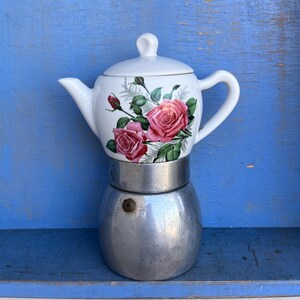 May include: A stovetop coffee maker featuring a white ceramic upper chamber decorated with a pink rose floral pattern and a matching lid. The lower section is constructed from brushed silver-toned metal and includes a small circular valve.