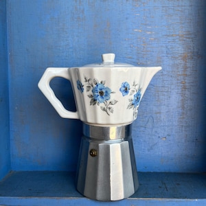 May include: A stovetop espresso maker featuring a white ceramic upper chamber with a fluted design and a blue floral pattern. The lower base is constructed from metallic silver aluminium and includes a small circular pressure release valve.
