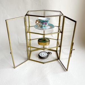 Puede incluir: Una vitrina hexagonal de vidrio y latón con dos puertas abiertas. En el interior, una taza y platillo, una pequeña lata y un reloj de bolsillo se exhiben en estantes de vidrio. La vitrina tiene una estética vintage.