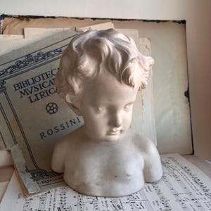 以下が含まれることがあります： 巻き毛の子供の白い石膏の胸像。胸像は楽譜の上に置かれています。