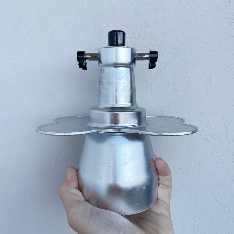 Op de afbeelding: Een zilveren metalen koffiezetapparaat met een zwarte knop bovenop en een bloemvormige basis. Het koffiezetapparaat wordt in een hand gehouden.