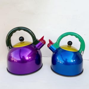 May include: Two colorful tea kettles. One is purple with a yellow lid and green handle, the other is blue with a yellow lid and green handle. Both have a black knob and red whistle.