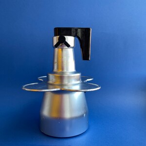 Op de afbeelding: Een zilveren aluminium koffiezetapparaat voor op het fornuis met een zwarte kunststof handgreep. Het koffiezetapparaat heeft een drietandvormige basis.