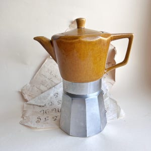 Puede incluir: Una cafetera de fogón con una parte superior de cerámica color amarillo mostaza que incluye tapa, asa y boquilla, unida a una base metálica de color plateado y forma octogonal. El diseño combina una parte superior de cerámica esmaltada.