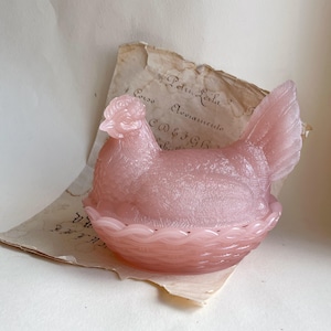 Może przedstawiać: Różowy szklany pojemnik w kształcie kury z podstawą w kształcie kosza i szczegółowymi piórami. Kura siedzi na kawałku postarzałego papieru z wyblakłym pismem. Dekoracyjny przedmiot kuchenny.